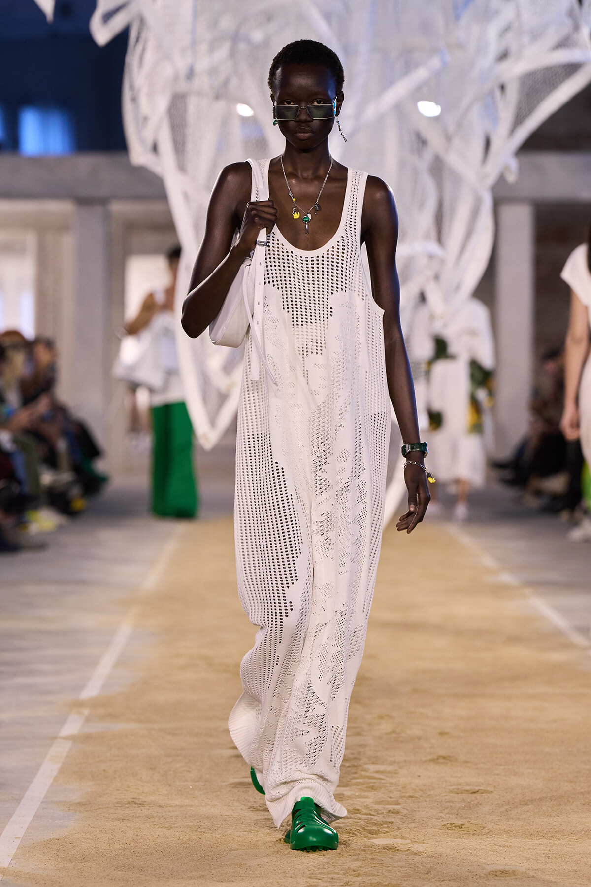 Model walks a runway in a white crochet maxi dress with a white shoulder bag and bright green shoes.