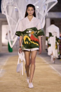 Model walking a fashion runway in a white oversized shirt paired with bold camouflage shorts and a white crossbody bag.