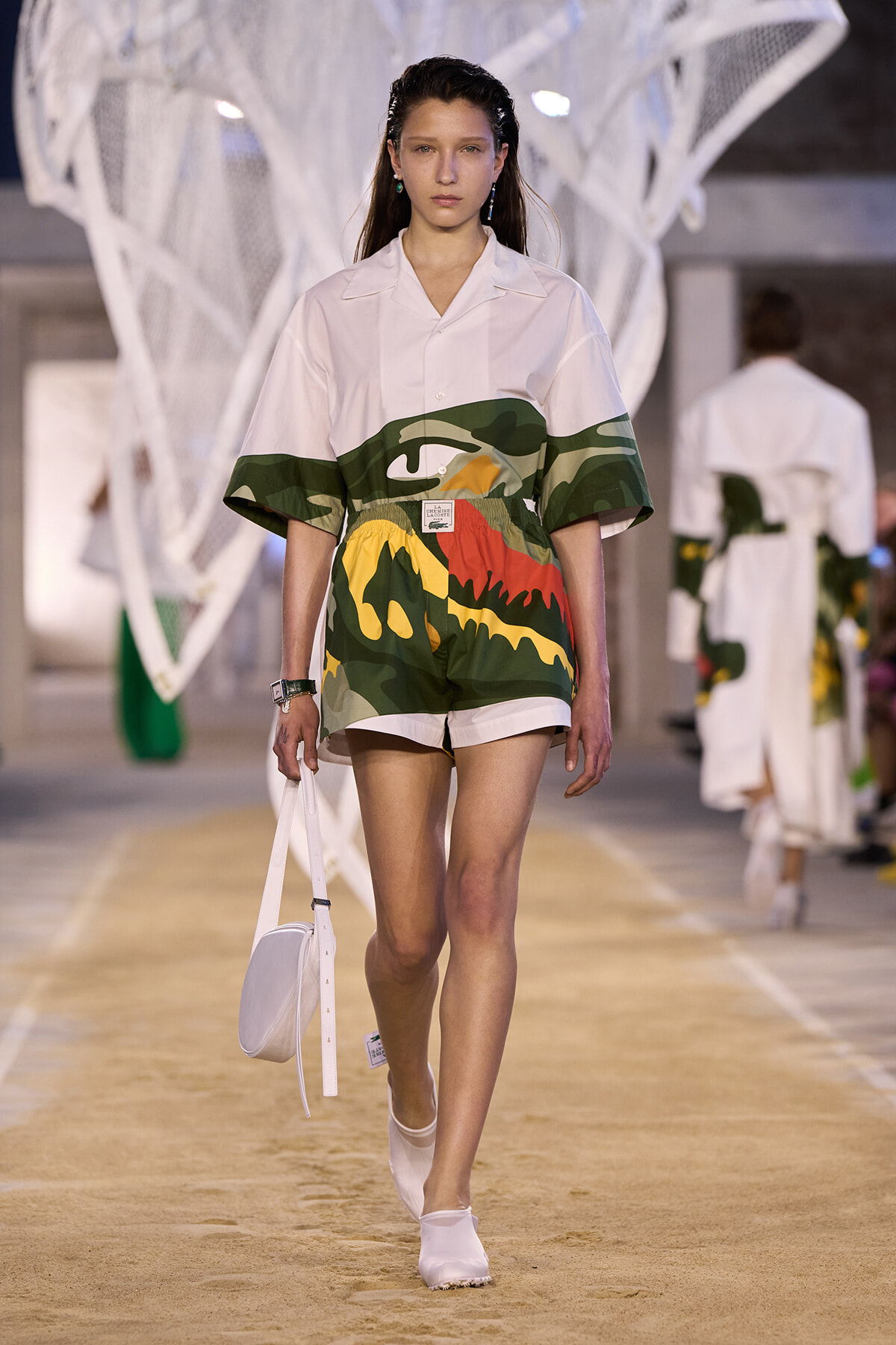 Model walking a fashion runway in a white oversized shirt paired with bold camouflage shorts and a white crossbody bag.