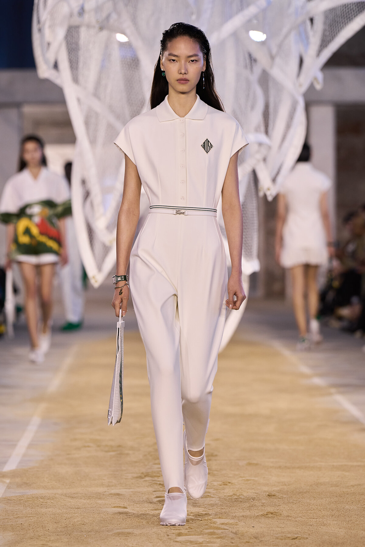 Model walks a white jumpsuit with a slim belt down a sandy runway, holding a white clutch; diamond logo on the chest and minimalist styling overall.