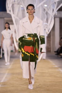 Runway model wearing a white oversized coat with a bold camo-print wrap skirt and white ankle boots, walking down a fashion show runway.