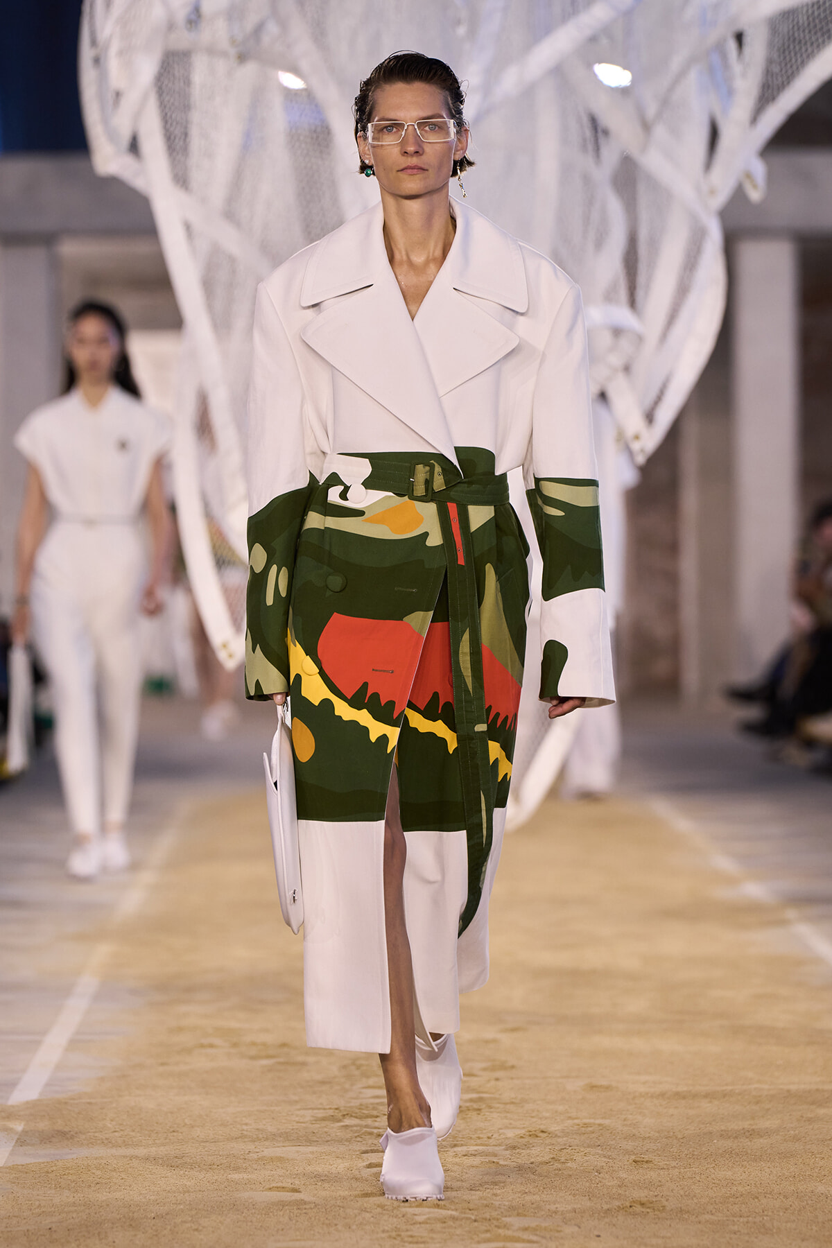 Runway model wearing a white oversized coat with a bold camo-print wrap skirt and white ankle boots, walking down a fashion show runway.
