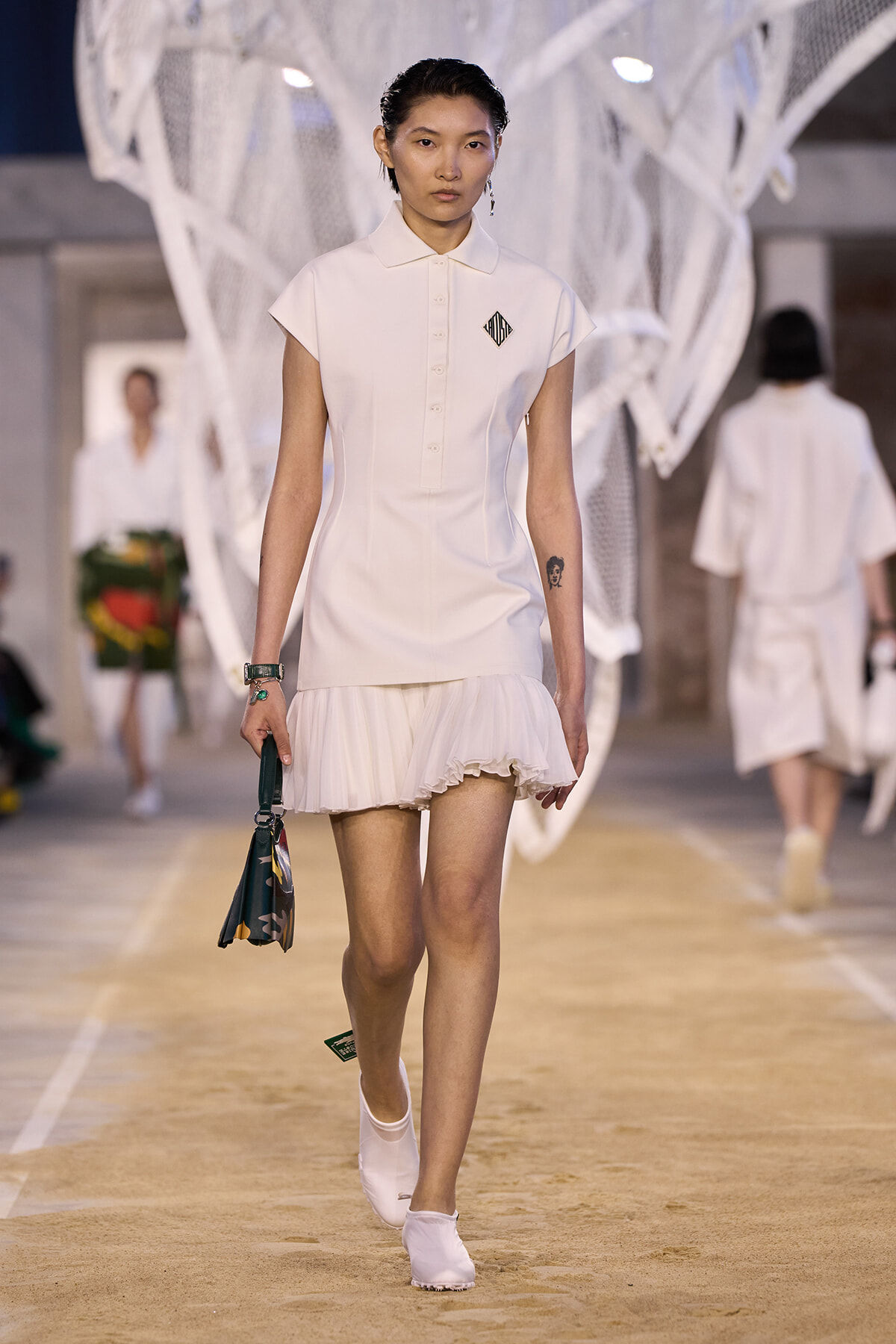 Model walks a fashion show wearing a white polo-style dress with a pleated ruffled mini skirt while carrying a patterned handbag.