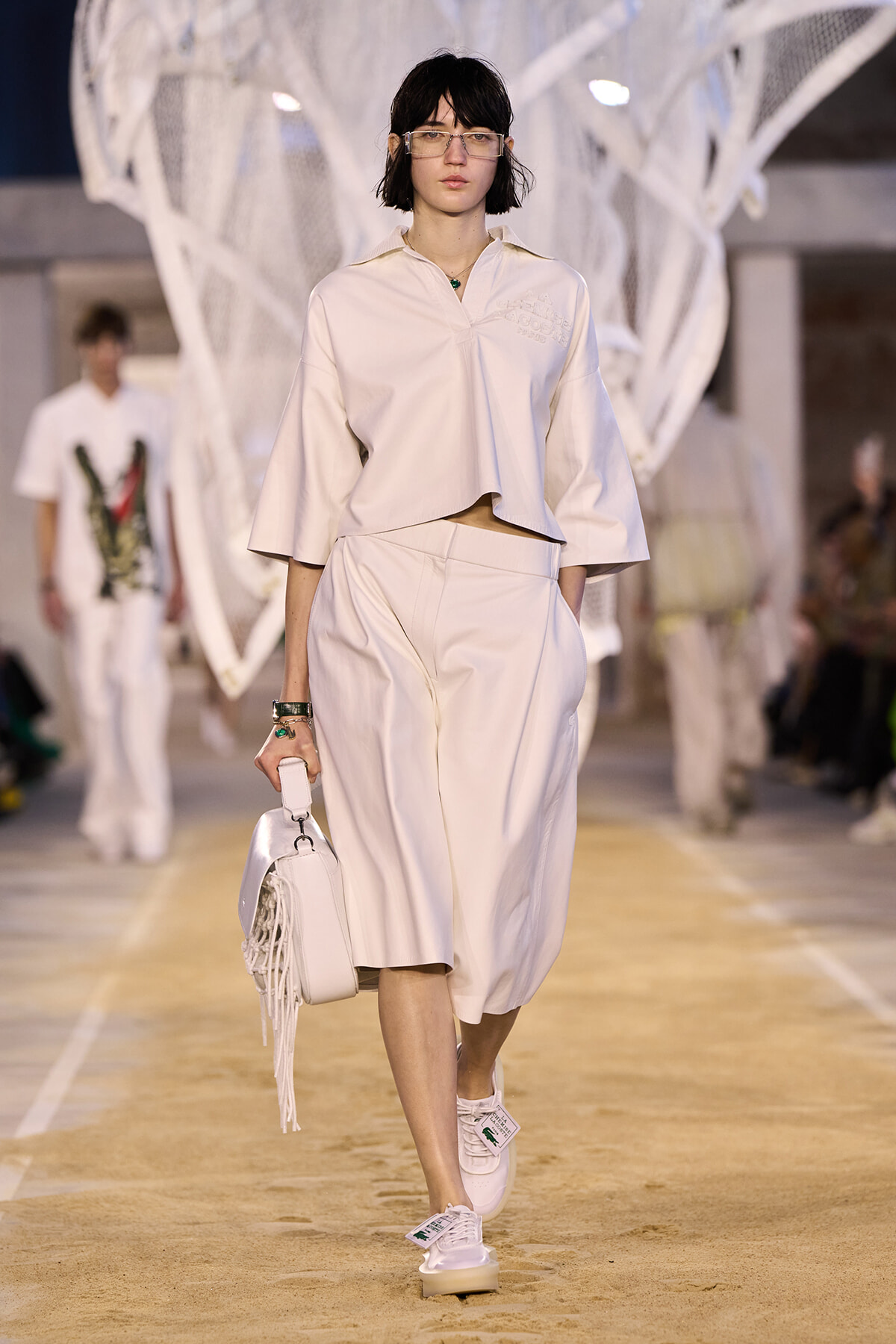 Model walks the runway in an all-white cropped top and wide-leg skirt, carrying a white fringed bag and wearing clear-framed glasses.