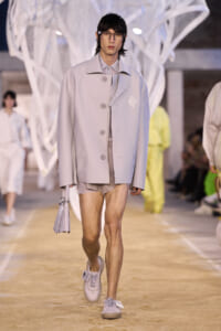 Male model walks a runway in a pale gray oversized coat, beige shorts, white sneakers, and clear glasses, carrying a small lavender bag.