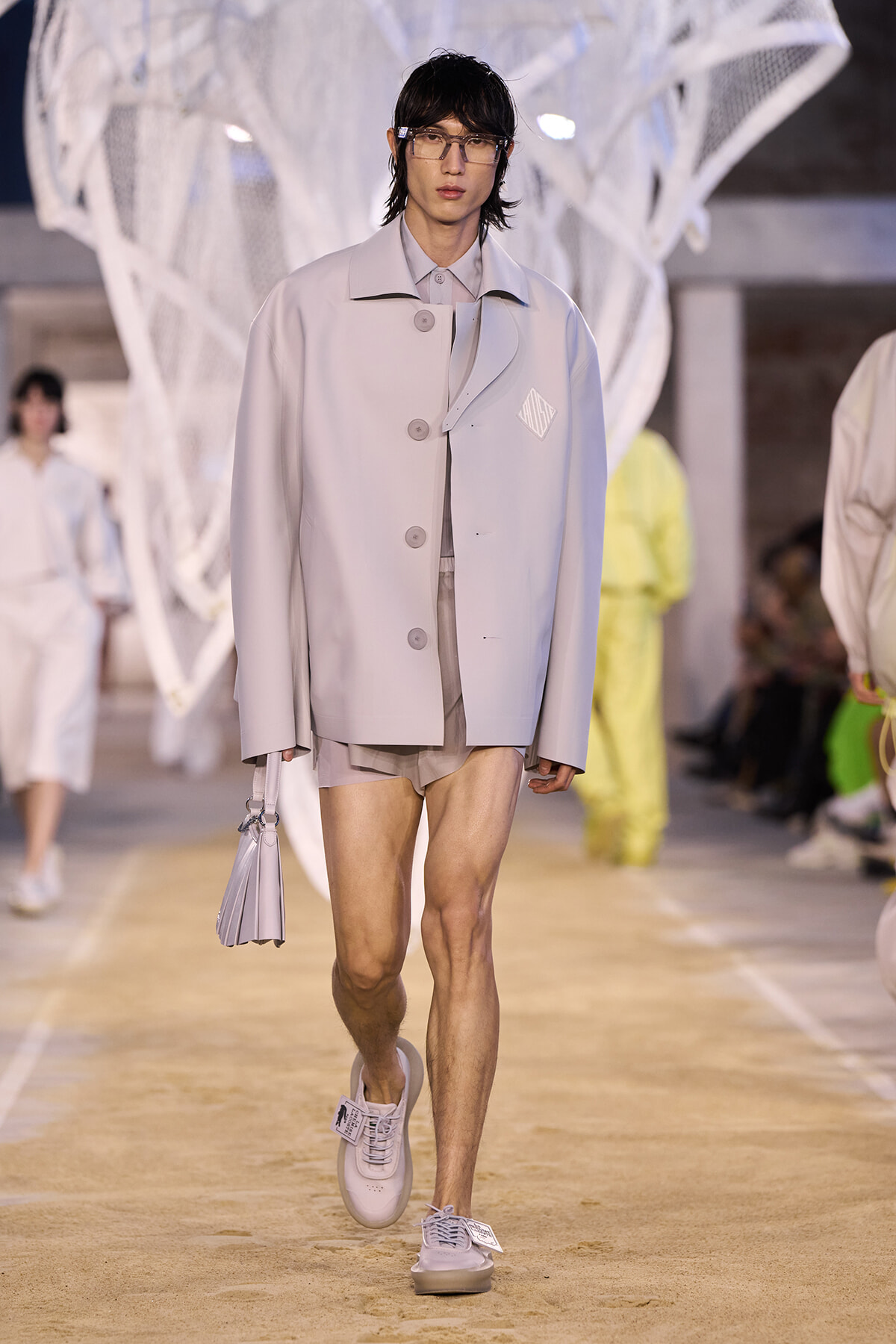 Male model walks a runway in a pale gray oversized coat, beige shorts, white sneakers, and clear glasses, carrying a small lavender bag.