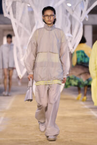 Model walks a runway in a beige oversized windbreaker and matching pants, carrying a pale handbag.