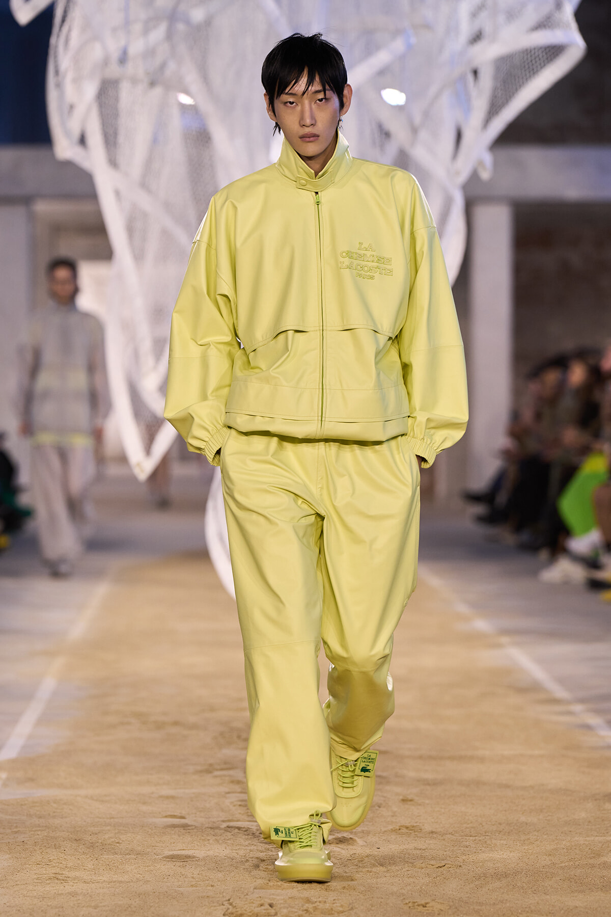 Male model in a pale yellow oversized zip-up jacket and matching pants walking a fashion runway.