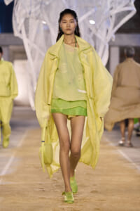 Model walks the runway in a sheer lime-green top, lime shorts, and a pale yellow oversized coat with matching shoes and bag.
