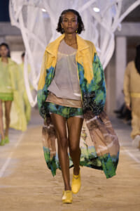 Model walking a runway in a yellow tropical-print coat over a beige top and matching shorts, with yellow footwear in a fashion show setting.