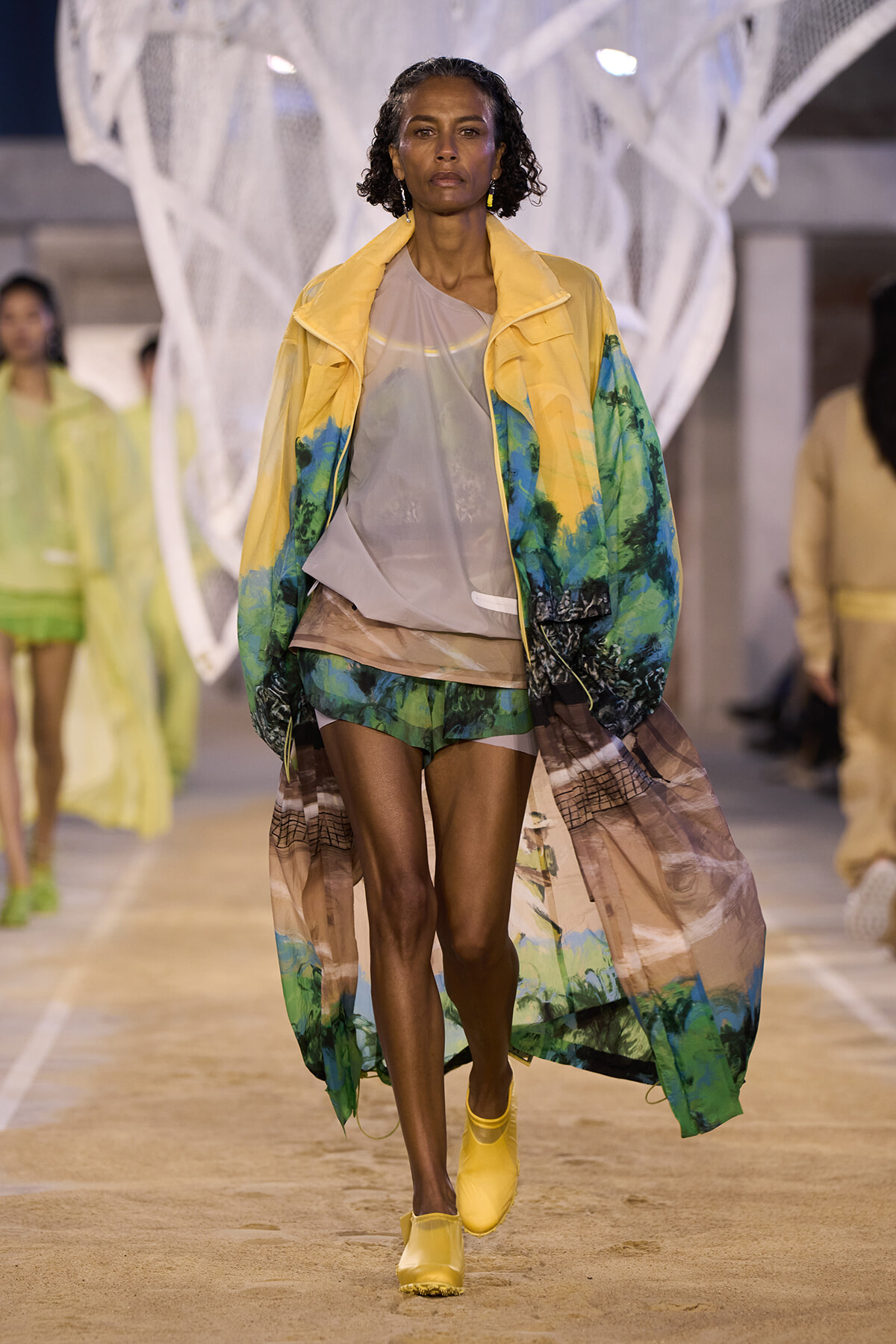 Model walking a runway in a yellow tropical-print coat over a beige top and matching shorts, with yellow footwear in a fashion show setting.