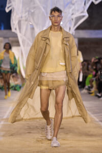 Male model walking a beige trench coat over a pale yellow top and layered shorts on a runway; futuristic, airy fashion scene with audience in background.