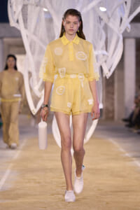Female model walking a fashion show in a pale yellow patchwork polo romper with white sneakers, holding a white water bottle.