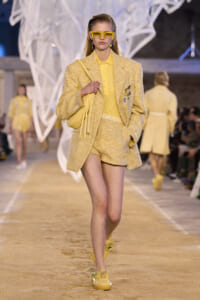 Model walking a fashion runway in a pale yellow tweed suit—blazer, high-waisted shorts, yellow bag, and oversized yellow sunglasses.