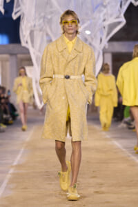 Model walks a runway in a pale yellow textured coat with a beige belt and matching yellow accessories on display heels/sneakers.