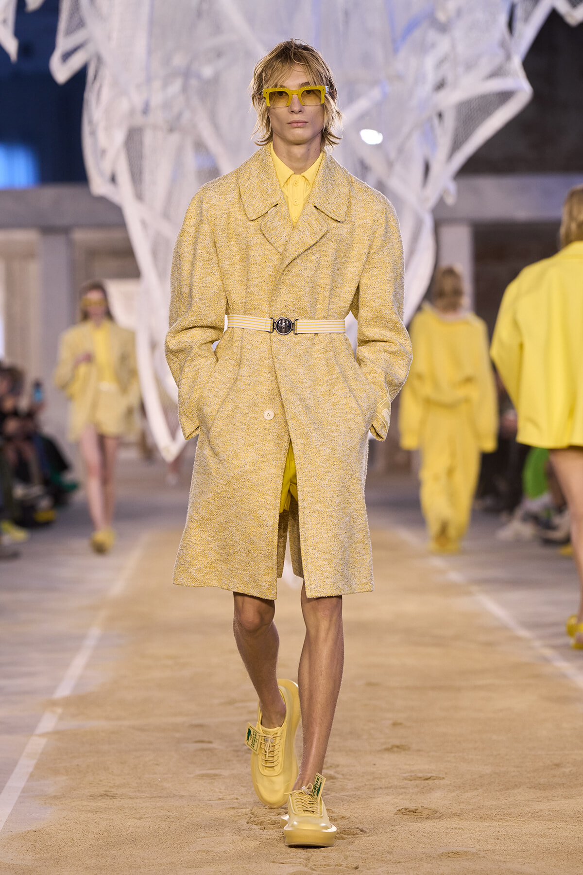 Model walks a runway in a pale yellow textured coat with a beige belt and matching yellow accessories on display heels/sneakers.