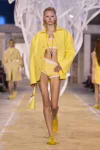 Model walking the runway in a bright yellow coat over a yellow knit bikini set, yellow sandals, and a small yellow handbag.
