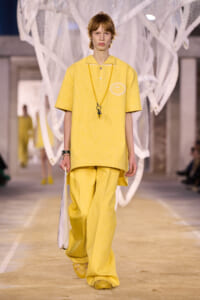 Model wearing a loose yellow polo and wide-leg pants on a runway, holding a white bag and wearing a long beaded necklace.
