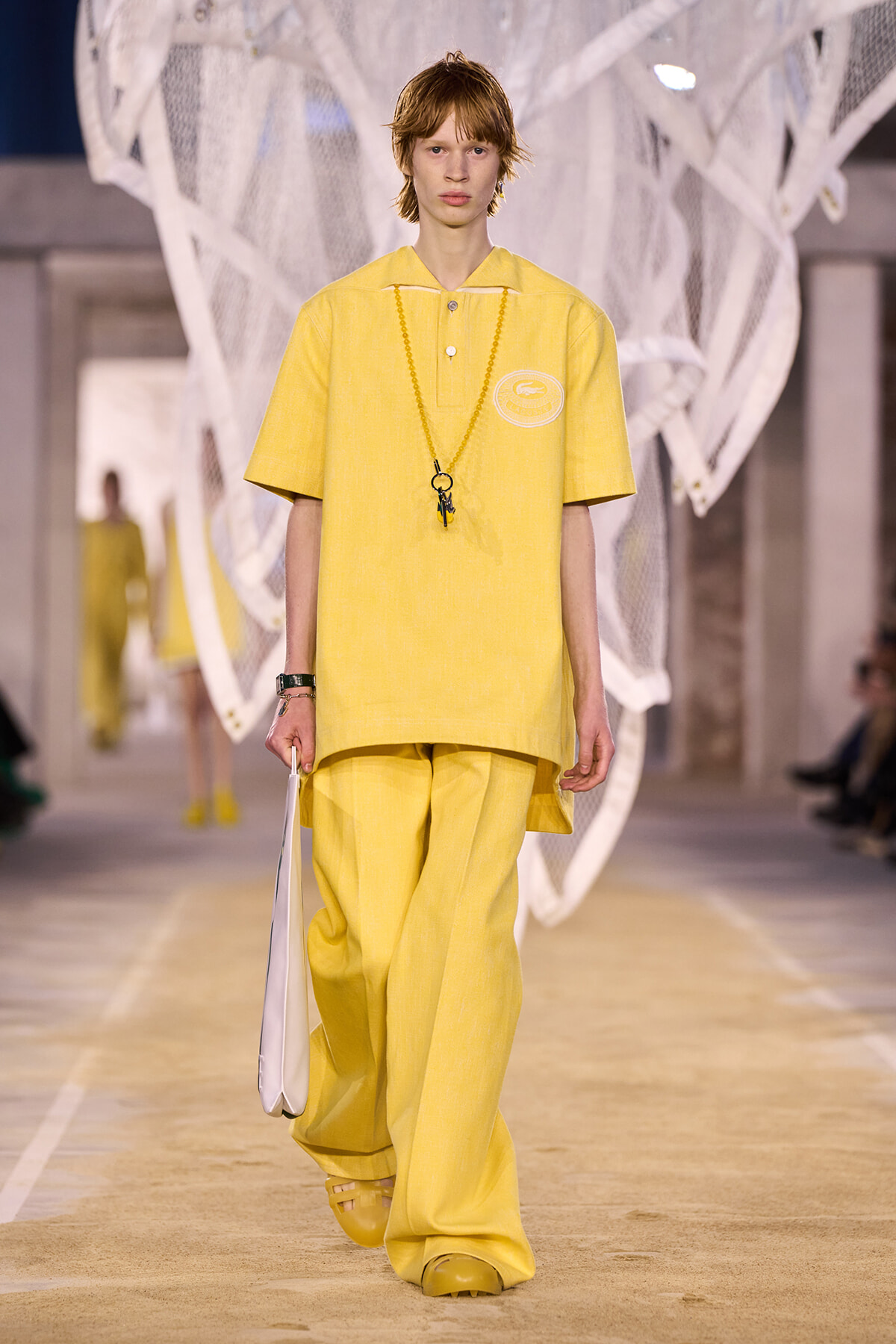 Model wearing a loose yellow polo and wide-leg pants on a runway, holding a white bag and wearing a long beaded necklace.