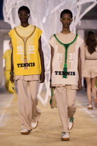 Two models walk a runway wearing oversized knit vests with a tennis motif and the word 'TENNIS', beige wide pants, and sunglasses.