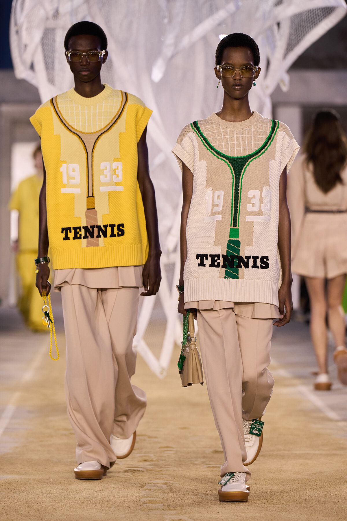 Two models walk a runway wearing oversized knit vests with a tennis motif and the word 'TENNIS', beige wide pants, and sunglasses.