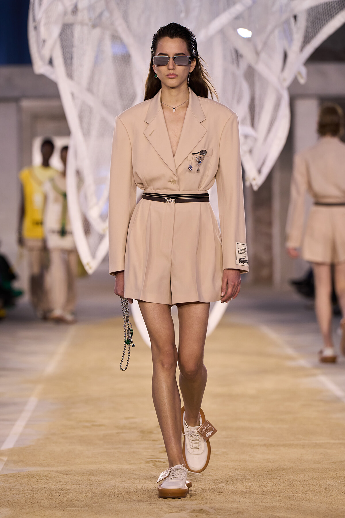 Runway model in a beige belted blazer romper with sunglasses and a small chain bag, walking a fashion show runway.