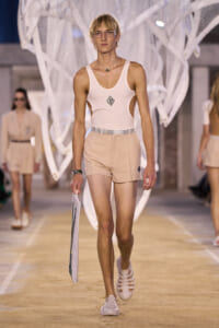 Male runway model walks in a light pink sleeveless top and beige shorts, holding a white poster board during a fashion show.