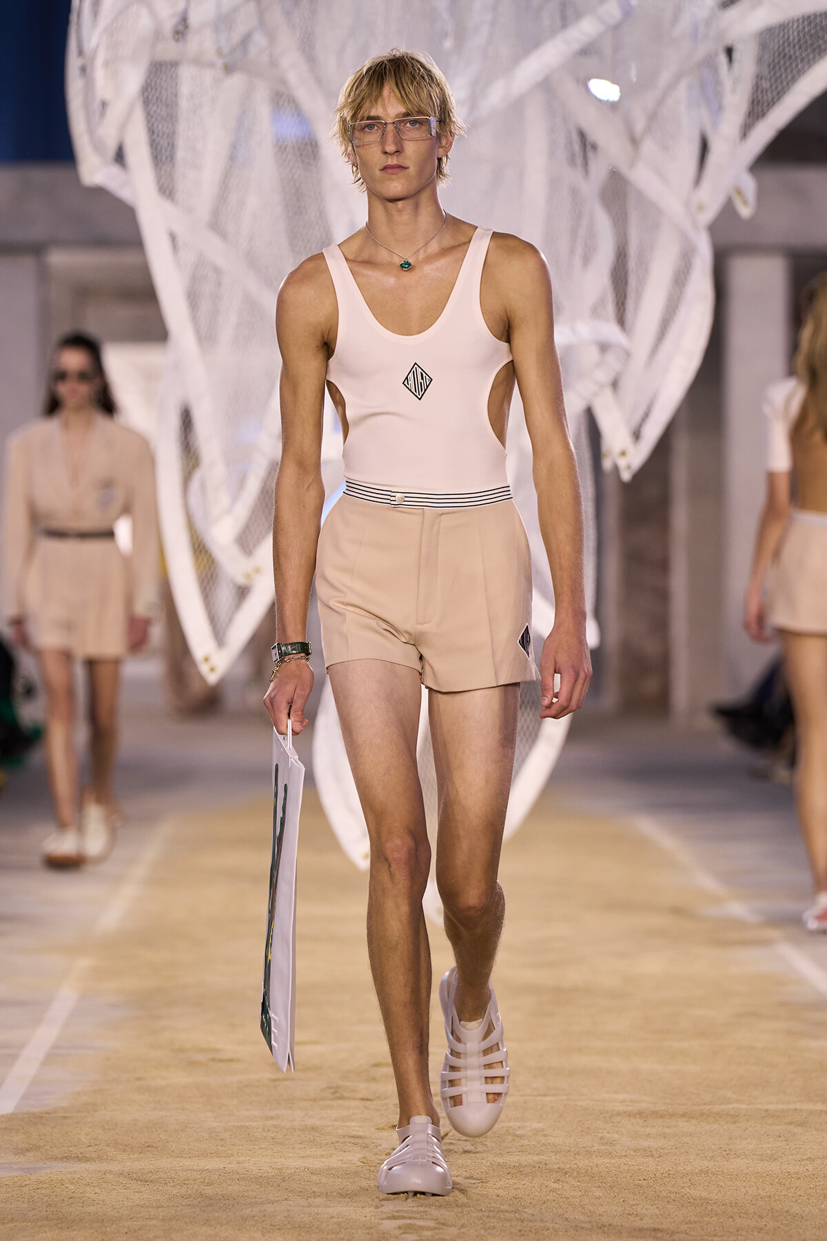 Male runway model walks in a light pink sleeveless top and beige shorts, holding a white poster board during a fashion show.