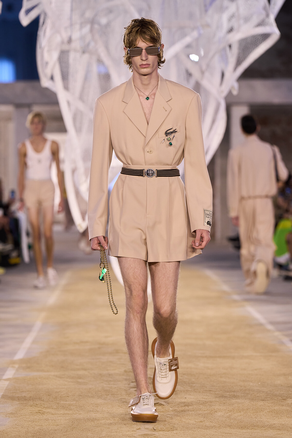 Male model in a beige tailored blazer romper with belt, sunglasses, and a green pendant, walking a runway holding a chain accessory.