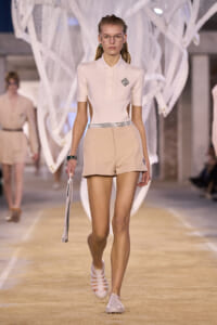 Model walks a fashion runway wearing a pale pink polo top with cutouts at the sides and beige shorts, white sandals, and clear glasses.