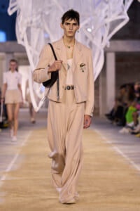 Male model walks a runway in a beige suit with a cropped blazer, wide trousers, and a black shoulder bag. Emerald jewelry accents visible.