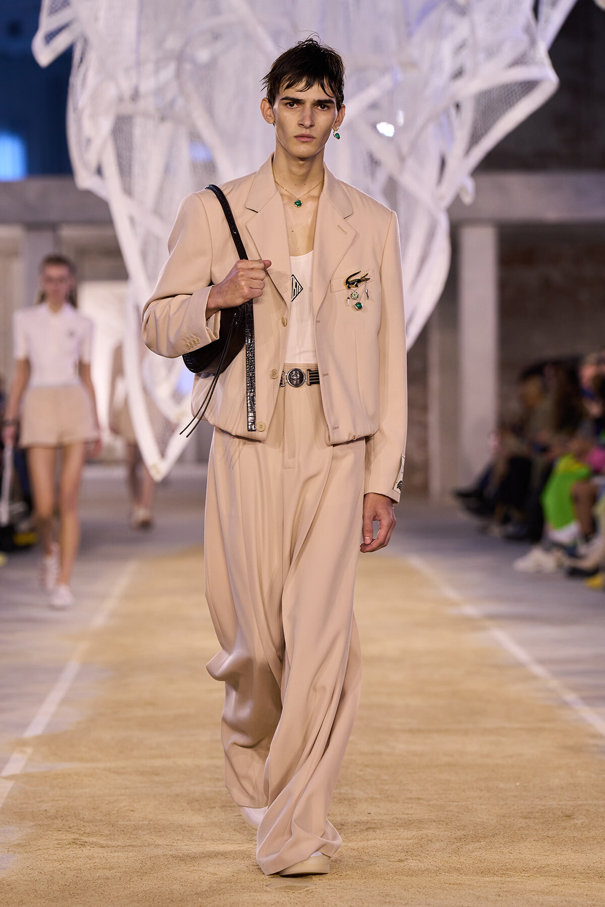 Male model walks a runway in a beige suit with a cropped blazer, wide trousers, and a black shoulder bag. Emerald jewelry accents visible.
