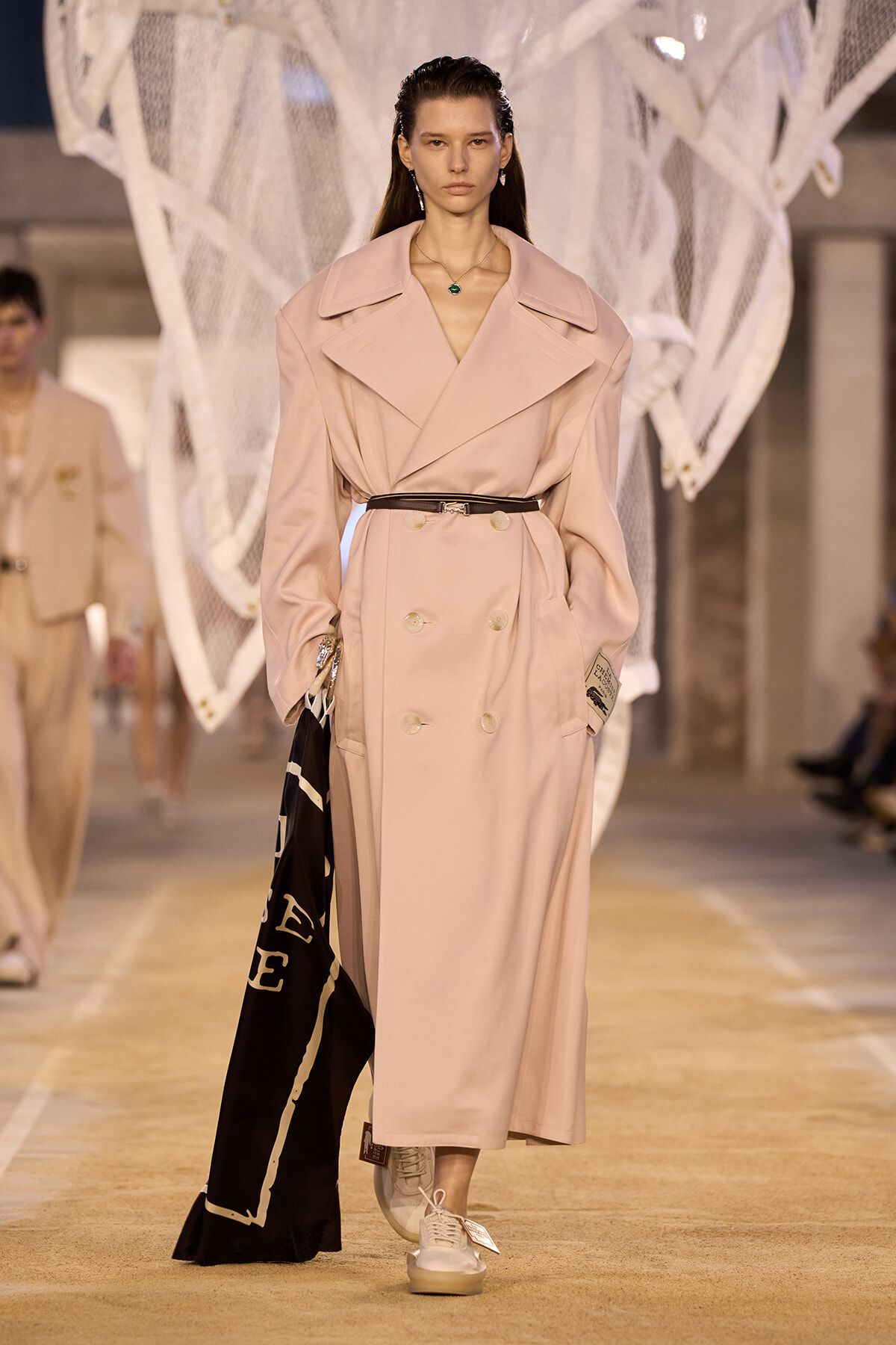 Model on a runway wearing a pale pink double-breasted trench coat with a belt, white sneakers, and a black scarf held at her side.