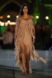 Model walking on a runway at night in a beige, fringe-tragged dress with thin straps and strappy sandals.