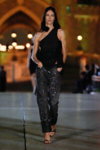 Model walking a nighttime runway in a black one-shoulder top and glittery cropped pants, sandals visible at the feet