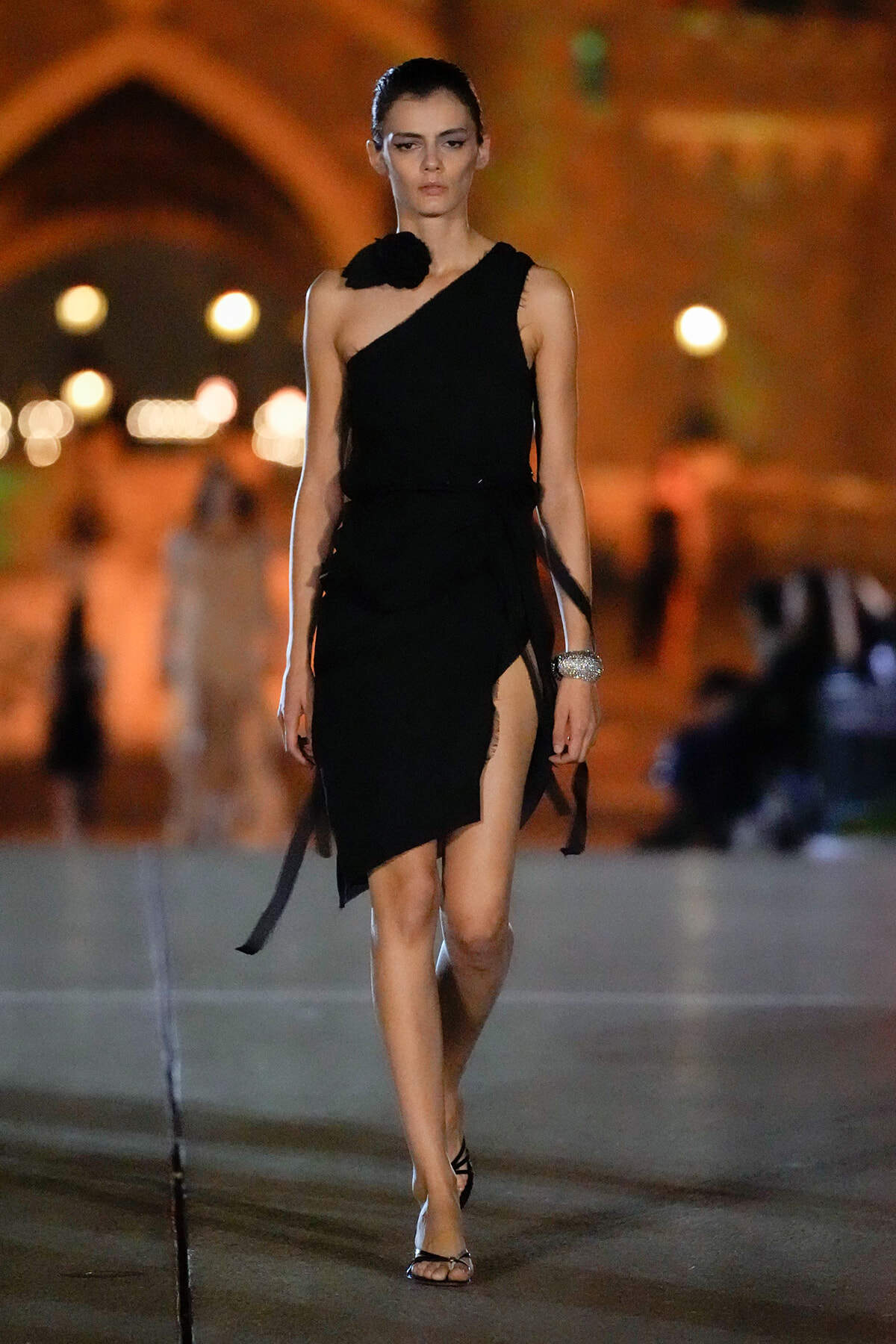 Female model walking a dimly lit runway in a black one-shoulder dress with a high slit, wearing strappy sandals.