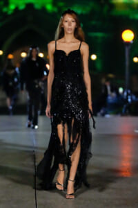 Model walking on a runway in a black sequined dress with thin straps, high-low slit, and strappy heels at night.