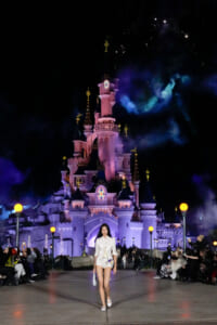 Fashion model in white outfit walking a runway at night, with a illuminated castle and cheering audience in the background.