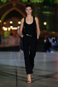Model walks down a dimly lit runway in a black sleeveless jumpsuit with a belt, hands in pockets, wearing strappy sandals; blurred city lights behind.