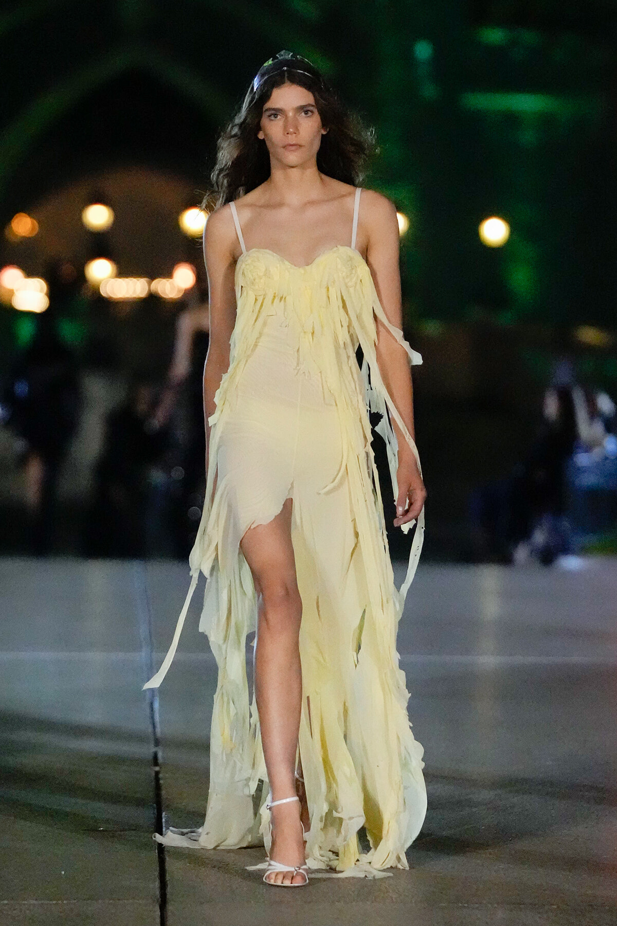 Female model walks a runway in a pale yellow fringed gown at night.