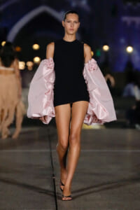 Model on a runway wearing a black sleeveless mini dress with oversized pale pink puff sleeves adorned with fabric roses.
