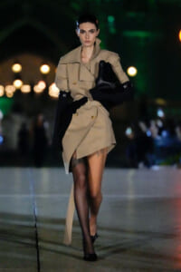 Female model walks a night runway in a tan trench coat, carrying a large black bag and wearing sheer black tights with black heels.