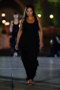 Woman walking toward the camera at night in a long black sleeveless dress and sandals, street lights in the background.