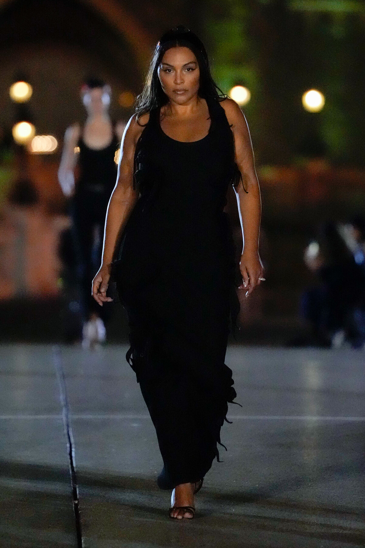Woman walking toward the camera at night in a long black sleeveless dress and sandals, street lights in the background.