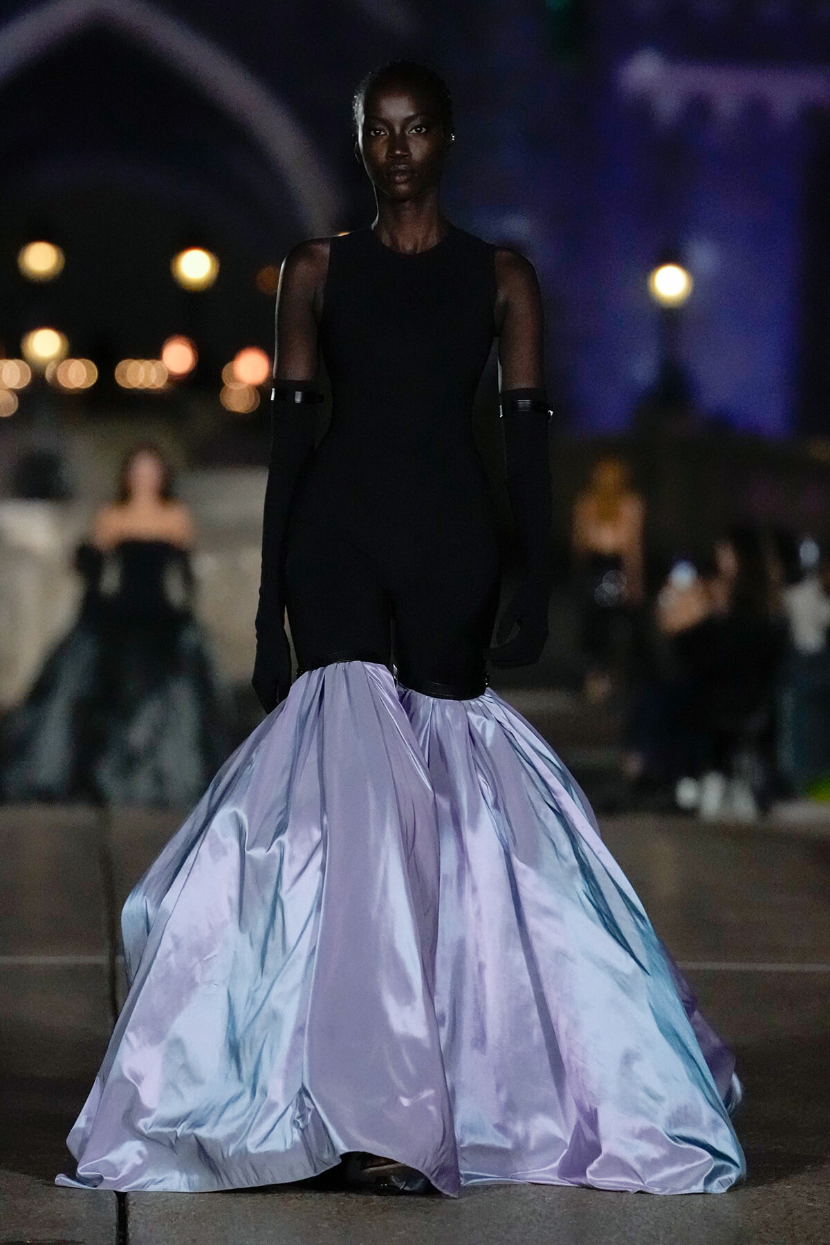 Model struts a fashion show in a black sleeveless bodysuit paired with a dramatic pale lavender, voluminous skirt at night.