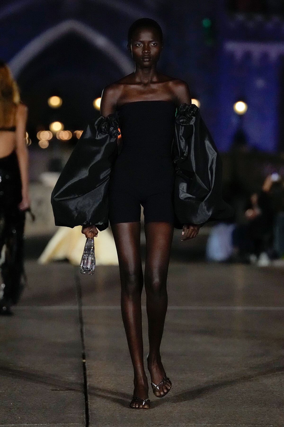 Model walking a night runway in a black off-shoulder dress with dramatic oversized sleeves, holding a water bottle