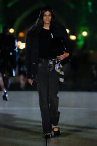 Model walking at night in an all-black ensemble: oversized jacket, black top, dark jeans with a striped scarf tied at the waist, black heels, and a black bag.