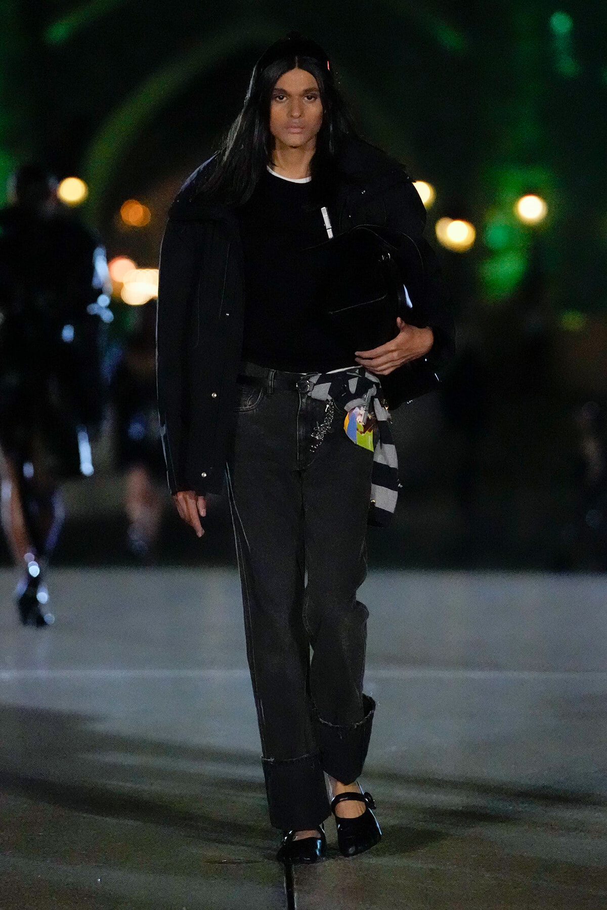 Model walking at night in an all-black ensemble: oversized jacket, black top, dark jeans with a striped scarf tied at the waist, black heels, and a black bag.