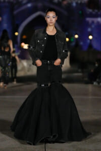 Model walking a runway in an all-black ensemble: black denim jacket over a black top and jeans with a dramatic, voluminous bell-shaped skirt.”
