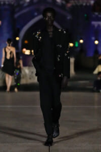 Model walking a night runway in a black embroidered suit with floral appliqués, moody blue lighting in background.